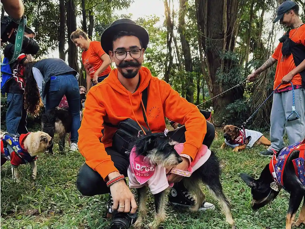 Man in orange hoodie and hat kneels with a dog in pink outfit at a park. Others with dogs gather around, creating a joyful community vibe.