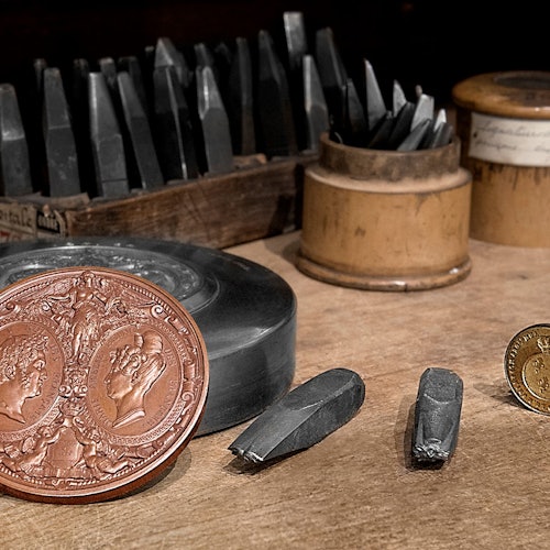 桌面上有雕刻工具的特写镜头、一枚放大的硬币，硬币上有两个面部轮廓，还有一枚金币和各种凿子。