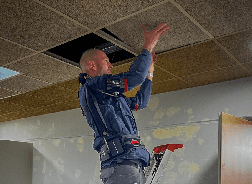 Technician wearing the Plum’ exoskeleton on a ladder to work on the ceiling, reducing fatigue and improving comfort at height.