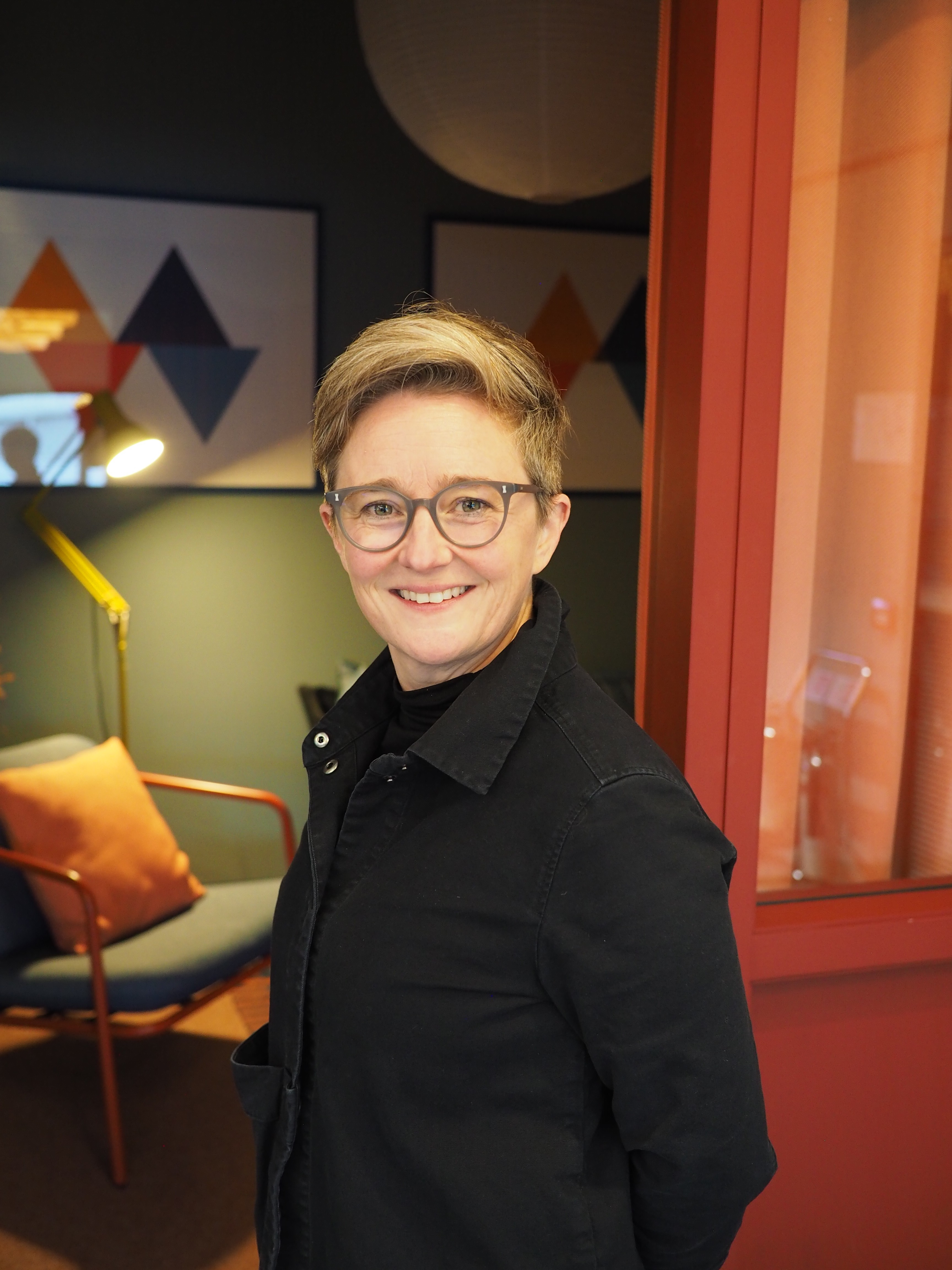 A person wearing a dark jacket stands in a warmly lit lounge-style room with geometric artwork on the wall, a lamp, and a chair with an orange cushion.