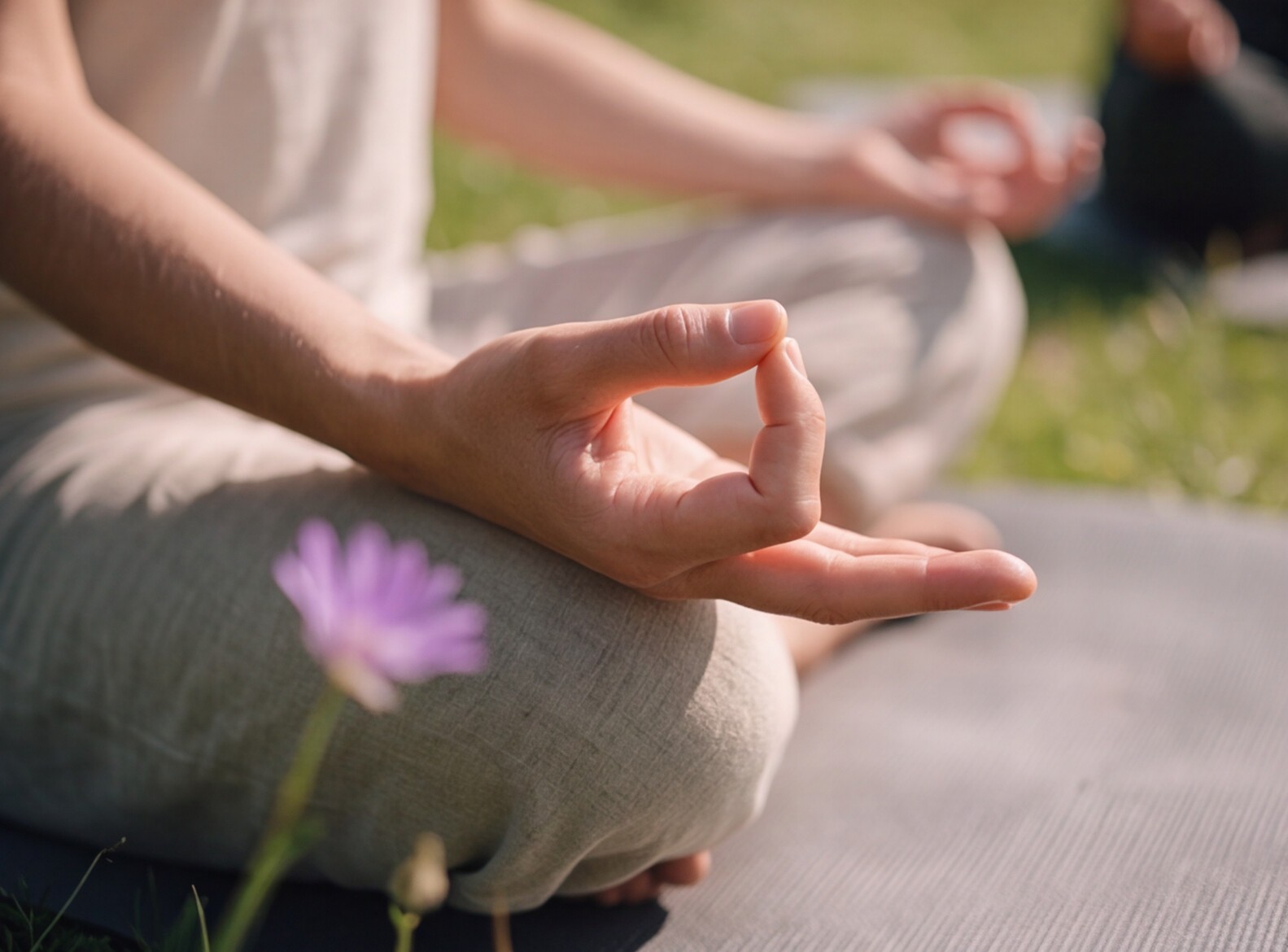 Close-up of a person sitting cross-legged outdoors in a meditation pose with hands in a mudra mudra gesture, next to a purple flower.
