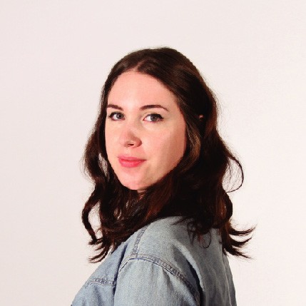 A woman with shoulder-length dark brown hair is shown from the back, wearing a light blue denim jacket. Her head is turned slightly to her left, and she looks directly at the camera with a neutral expression. The background is a plain light beige or off-white color.