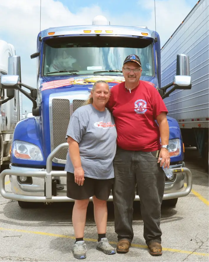 A couple who drives together standing in front of their blue truck, putting their arms around each other, and smiling