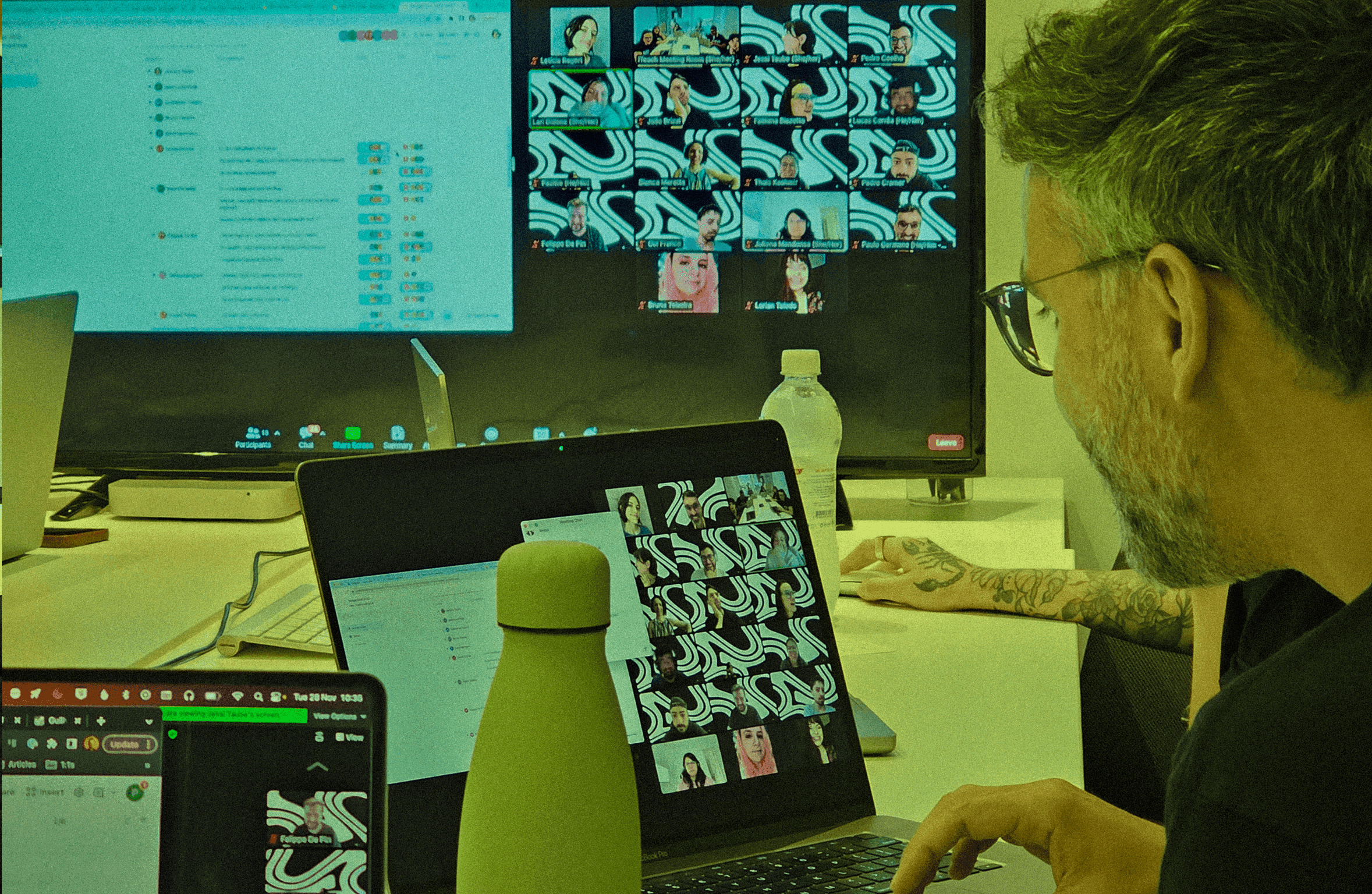 A professional working at a desk setup featuring a laptop and an external monitor displaying a large virtual meeting gallery.