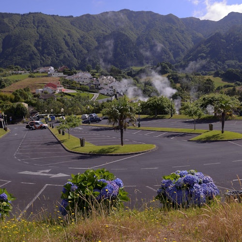 موقف سيارات مليء بالسيارات، محاط بالخضرة والزهور، يؤدي إلى منازل سكنية مع جبال في الخلفية.