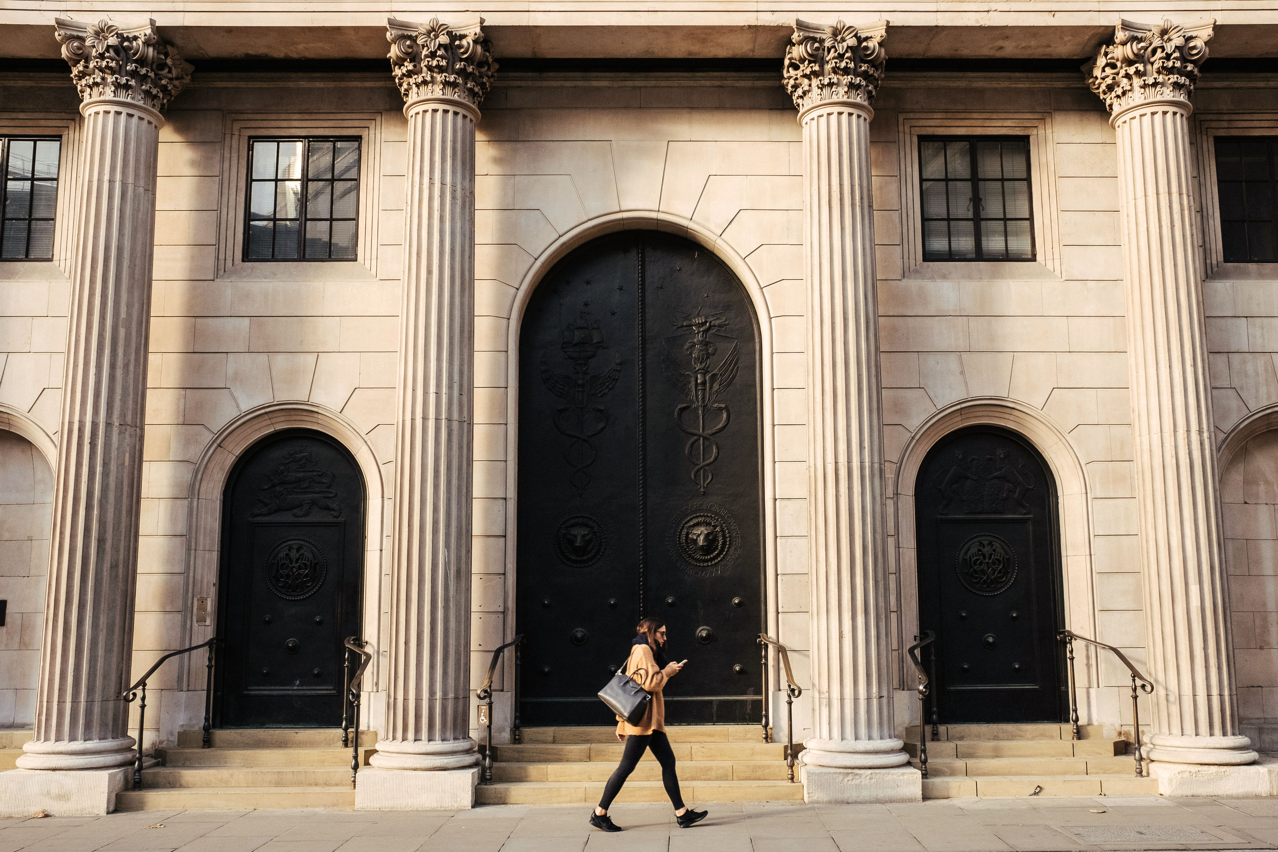 A person walks in front of large stone arches with dark doors, flanked by tall columns, in a historic building.