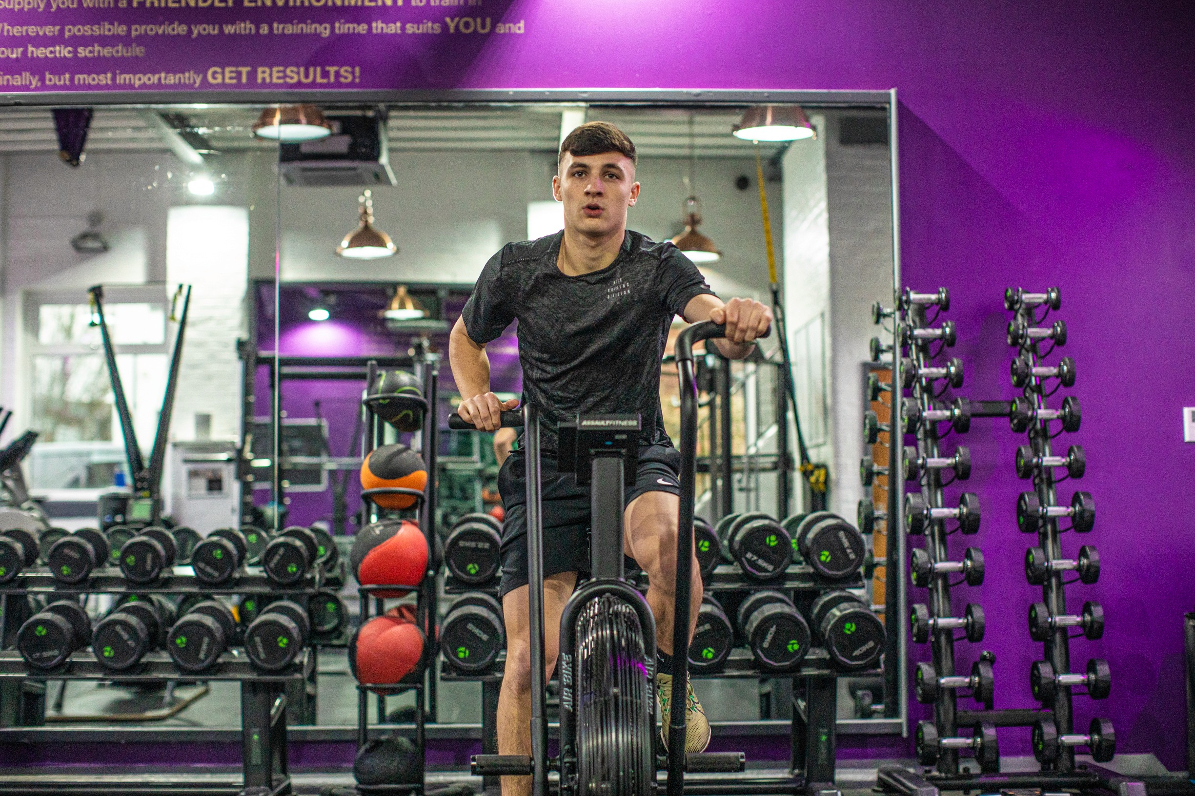 A young man is using a rowing machine in the gym, with weight plates and equipment in the background.