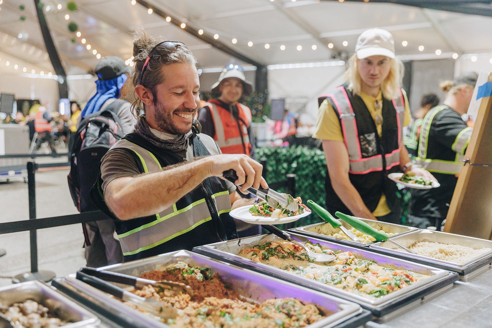 Two individuals at a food stall, one serving food and the other assisting, in a vibrant market setting.