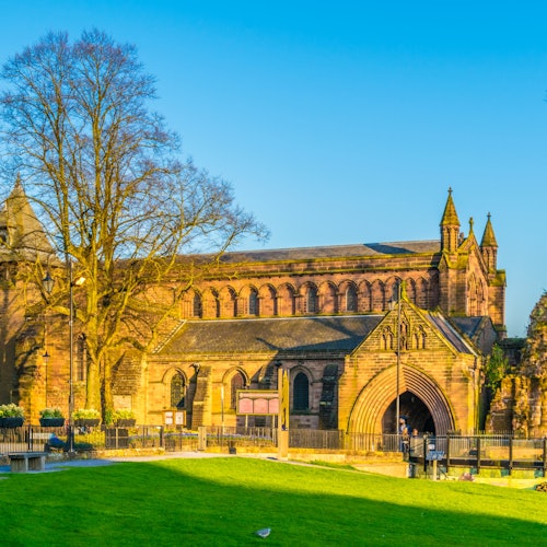 一座历史悠久的石砌建筑，拥有拱形入口、大窗户和塔楼，映衬在晴朗的蓝天下，周围环绕着绿草如茵的区域。