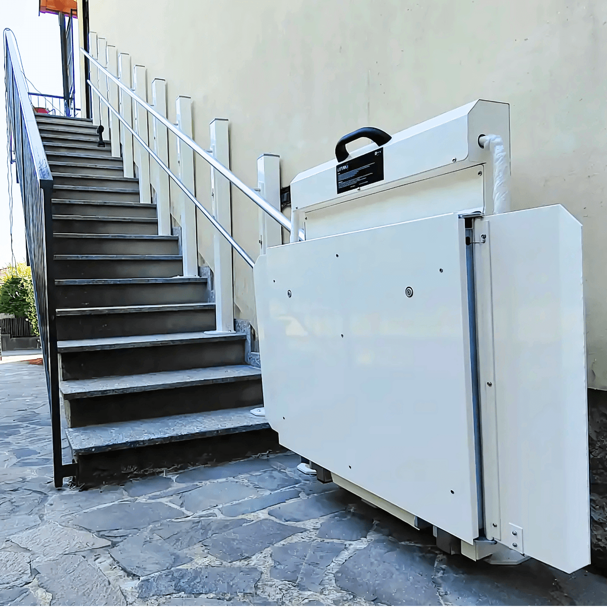 Incline platform stairlift in folded parked position at the base of an outdoor stone staircase — white folded platform and drive rail mounted against rendered wall with metal balustrade