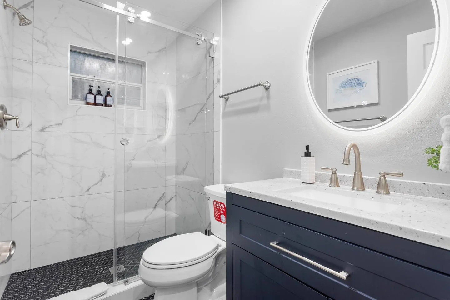 Hall bath with glass walk-in shower, lit round mirror and navy vanity at Sarasota Beach Bungalow, Bradenton, Florida