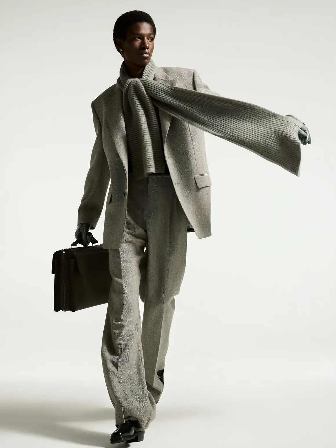 A model in a tailored gray suit with a matching scarf elegantly draped, holding a briefcase, striding confidently against a light neutral background, embodying high-end visuals for an AI production project in a fashion brand context.
