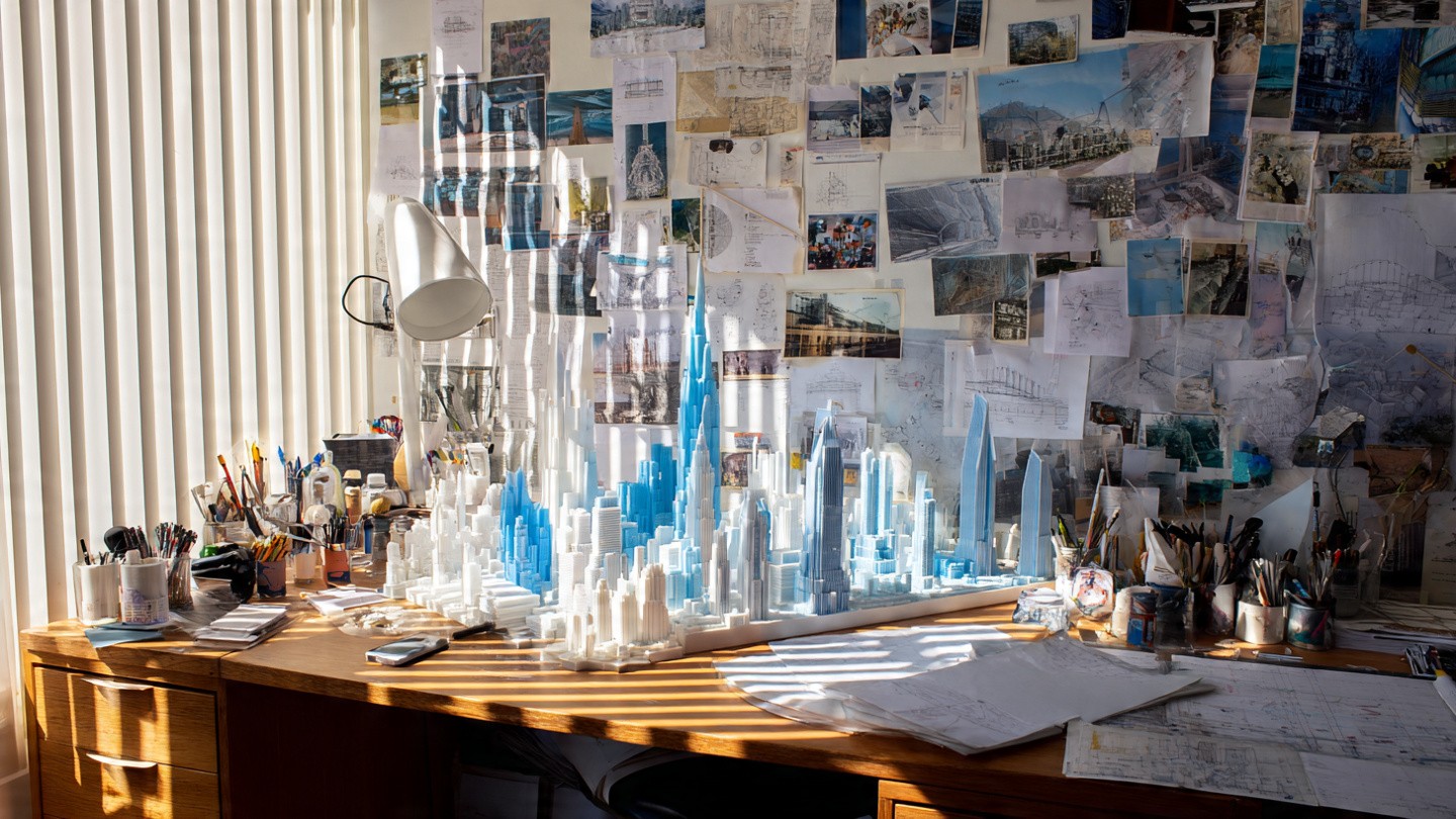 Architect's desk with a detailed model cityscape in white and blue; back wall is covered in various architectural sketches and images, lit by sunlight.