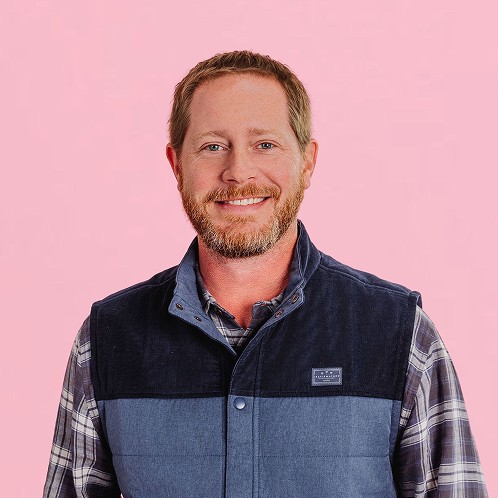 Smiling man with a beard wearing a plaid shirt and dark vest, standing against a pink background.