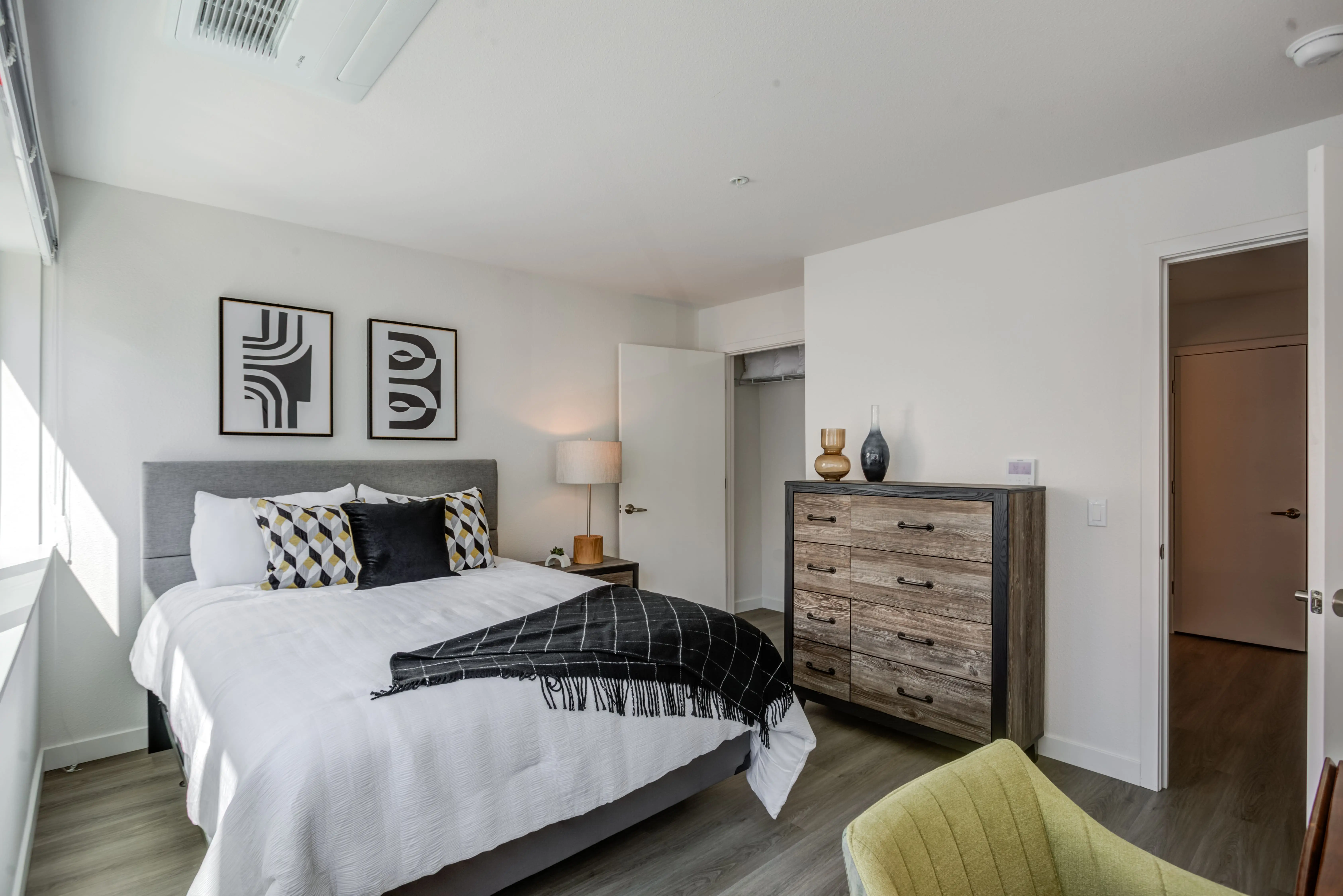 A bedroom in Midtown Sacramento is decorated in a modern style with a large bed featuring white bedding, black pillows, and a black throw. The room has a wooden dresser with decorative items, two abstract artworks on the wall, a nightstand with a lamp, and a green chair in the foreground—truly epitomizing modern living spaces.