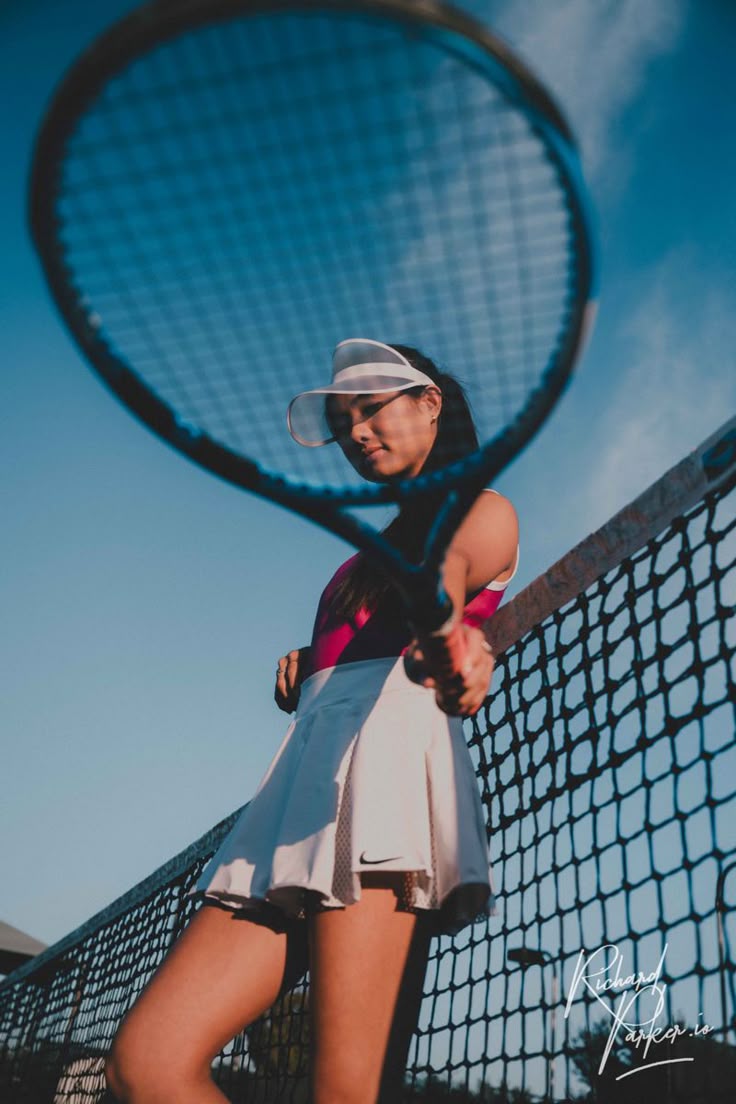 A female tennis player in a white visor and skirt holding a racket, posing on the court with a blue sky in the background.