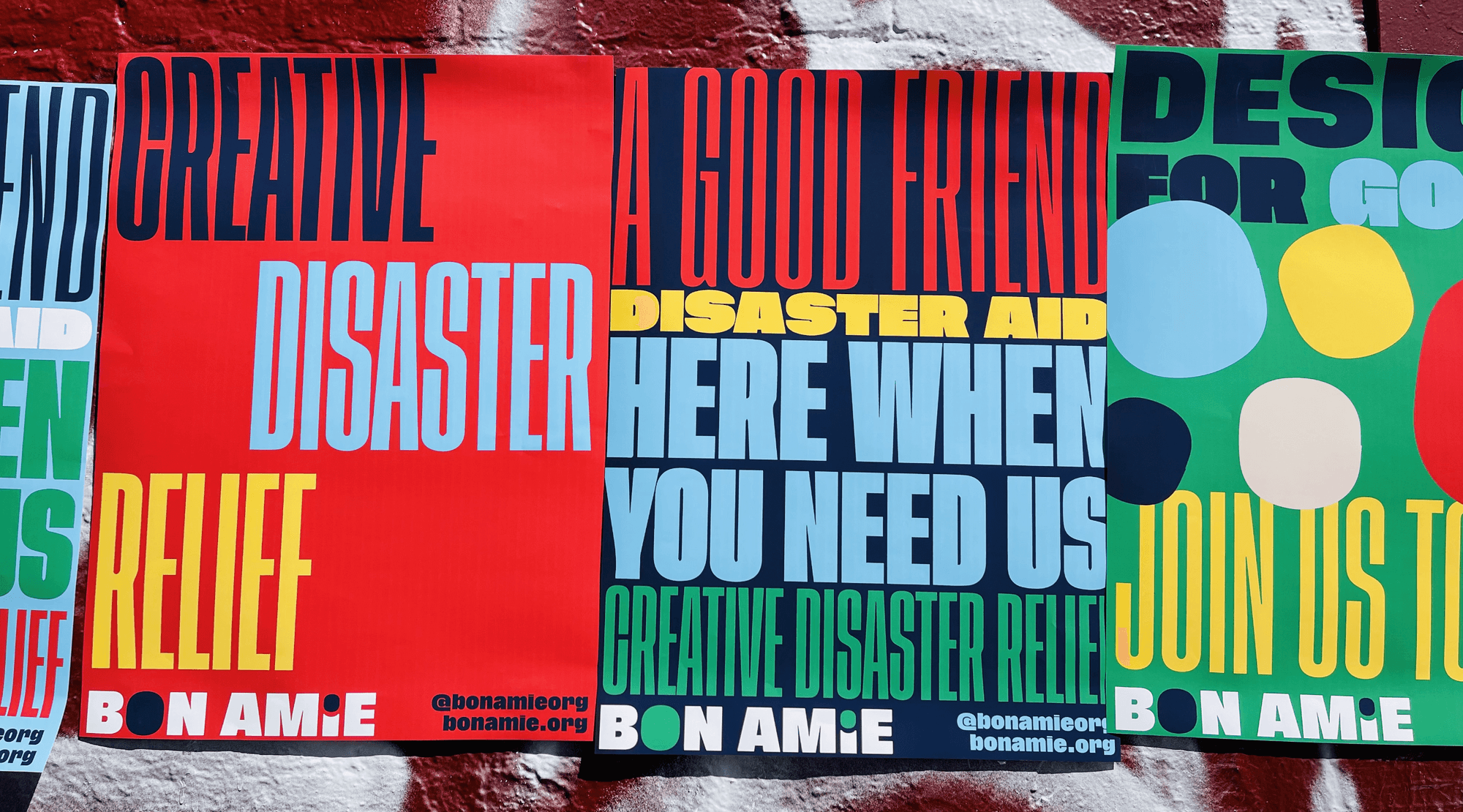 Three posters are displayed on a brick wall. The leftmost poster is red and features the words “CREATIVE DISASTER RELIEF” in bold black letters. The center poster is blue and displays “A GOOD FRIEND DISASTER AID HERE WHEN YOU NEED US” in blue letters. The rightmost poster is green and includes “DESIGN FOR GO JOIN US TO” in yellow letters. All posters have “BONAMIE” at the bottom.