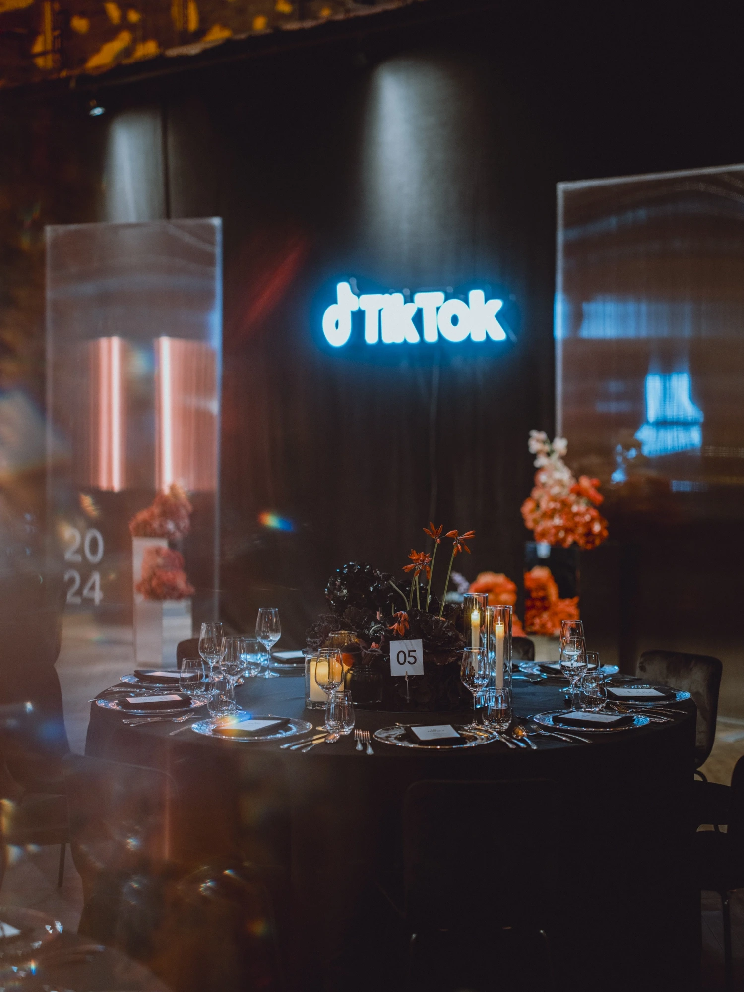 Black dinner table with candles, orange flowers and number card under the neon TikTok logo.