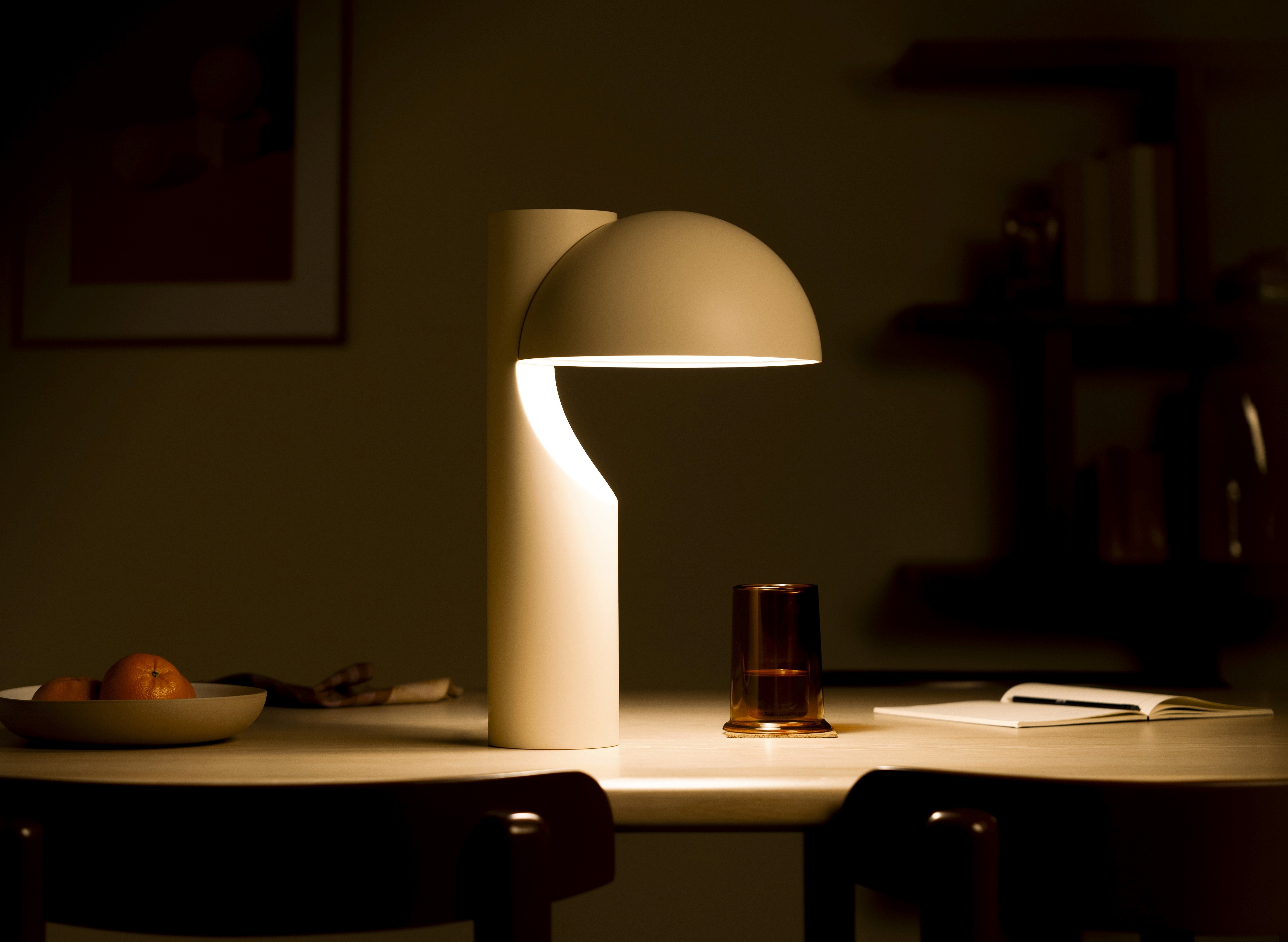 Cream table lamp with a dome shade, lit on a dining table at night beside an amber glass and an open notebook