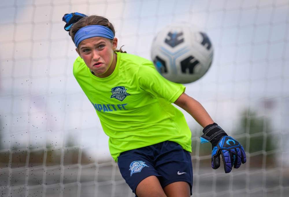A young female goalkeeper in neon green and blue unleashes a powerful kick, turf scattering around her as she stares down the ball with fierce determination.