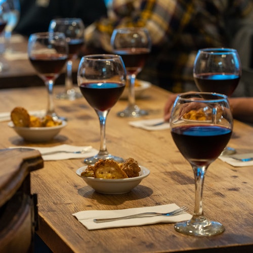 Several glasses of red wine and small bowls of appetizers on a wooden table with napkins and forks; blurred people in the background.