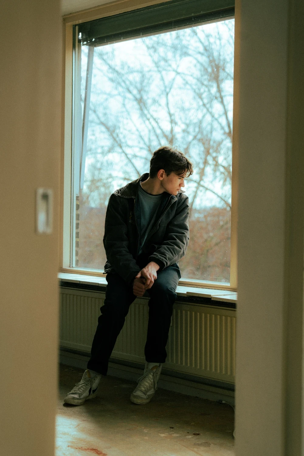Young man sitting alone by a large window, looking down in a quiet, reflective moment.