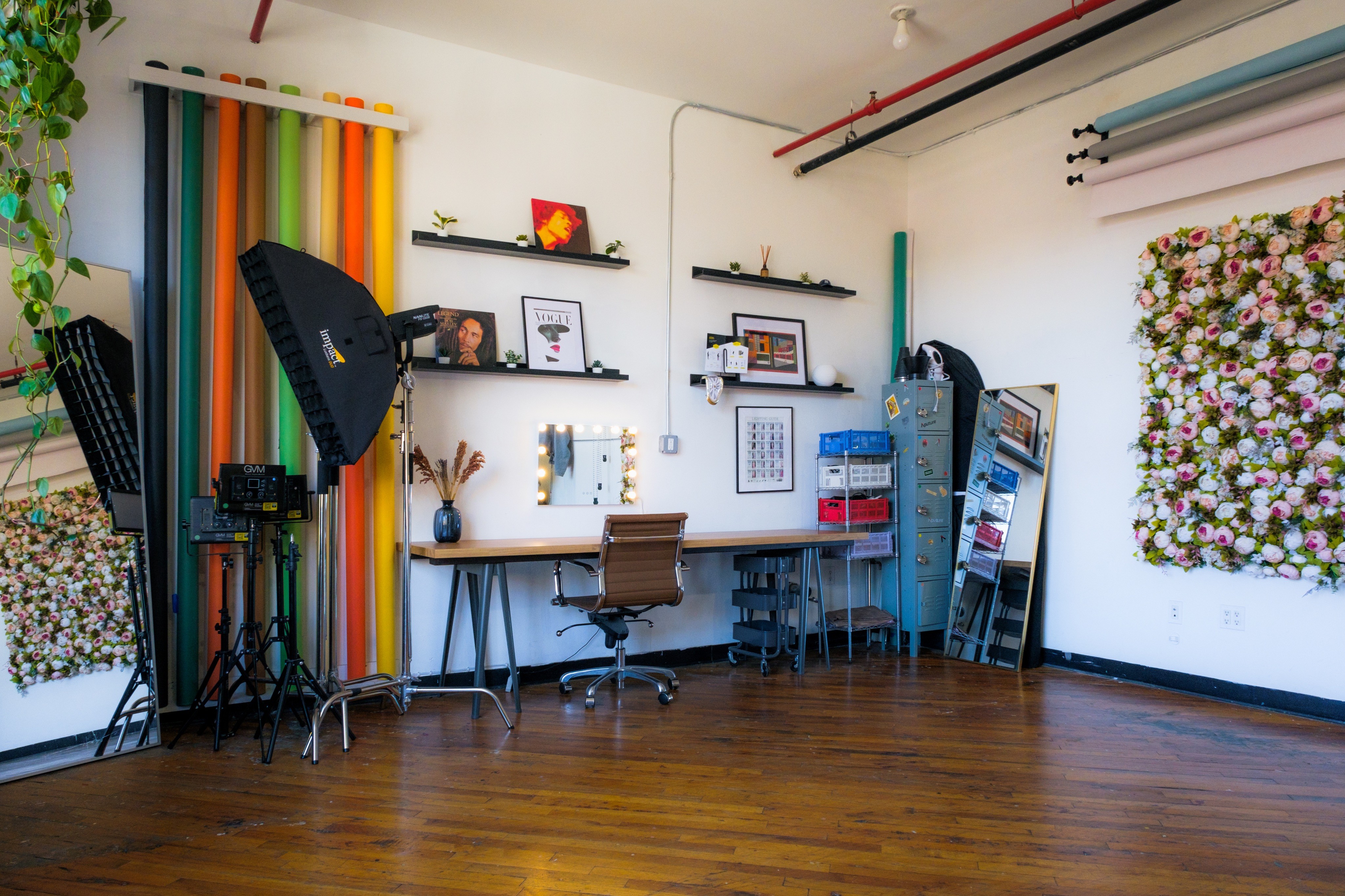 A daylight photography studio featuring a large wall of colorful backdrops, professional lighting equipment, and a workspace with shelves displaying decor and camera gear. A floral wall installation and a large mirror add artistic elements to the studio’s bright, versatile environment.