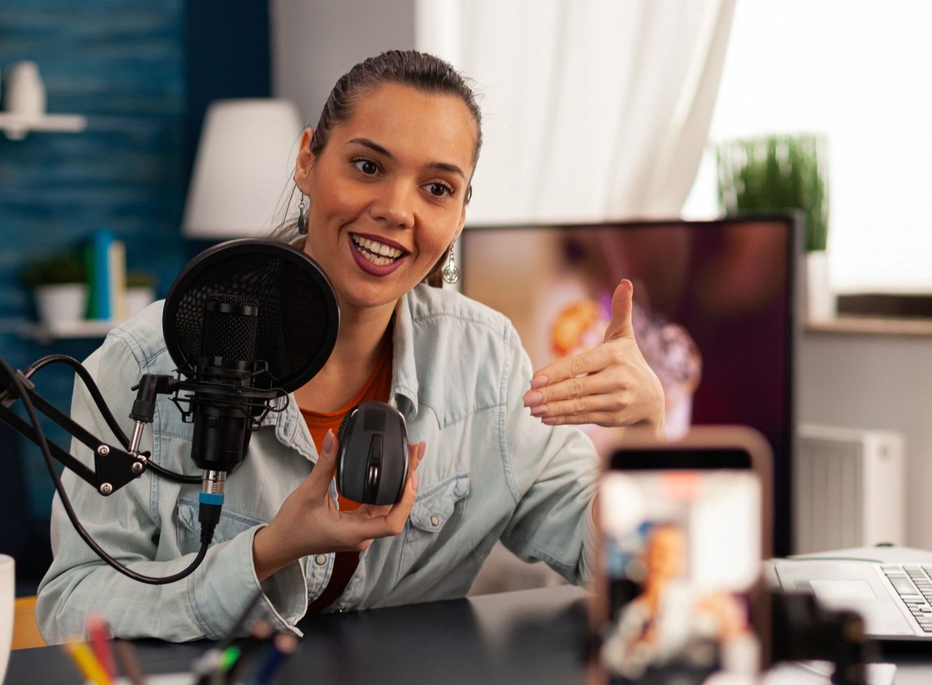 Una mujer sonriente grabando un podcast o video, hablando en un micrófono con filtro anti-pop, extendiendo una mano hacia la cámara como si estuviera explicando algo. 