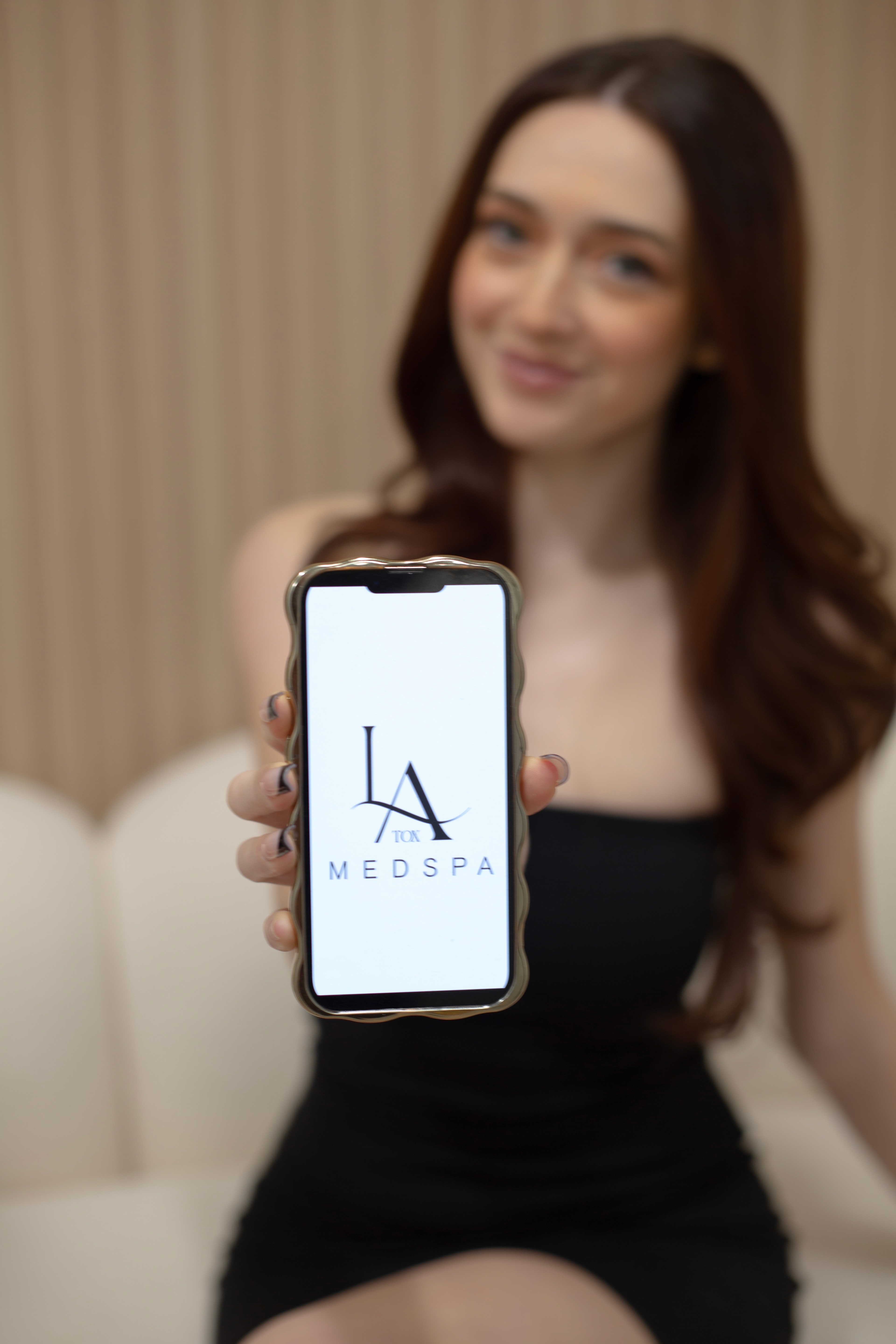 woman in Person holding a smartphone displaying the med spa logo on the screen.white scrub suit holding gray laptop computer