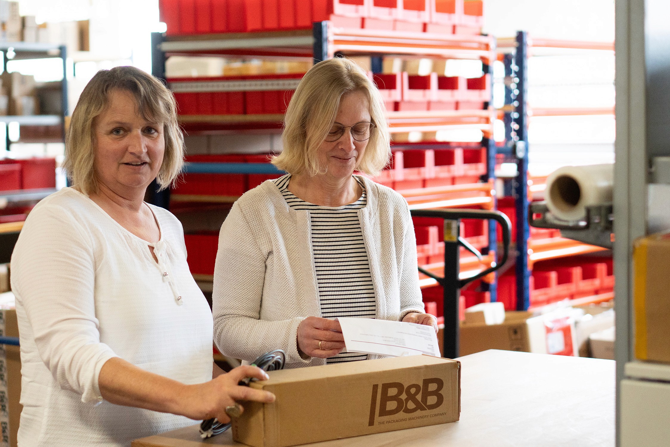 Zwei Frauen verpacken Pakete in Lager. Mitarbeiterinnen beim Versand mit B&B Logo.