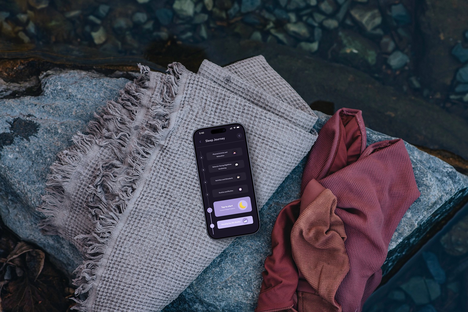 High-angle shot of a smartphone resting on textured grey and rust-colored fabrics placed on a rock by the water. The screen displays the Vialog "Sleep Journey" interface with its timeline and moon icon, blending digital well-being with a serene, natural environment.