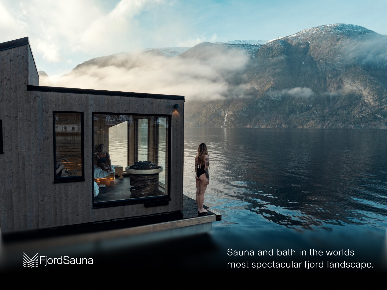 A person on a floating sauna looking at a beautiful fjord landscape with FjordSauna logo