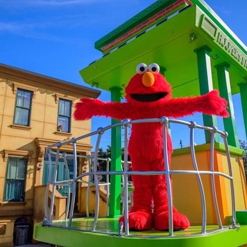 Елмо, голям червен куклен герой, стои с разперени ръце на зелен и жълт балкон до сграда под синьо небе.