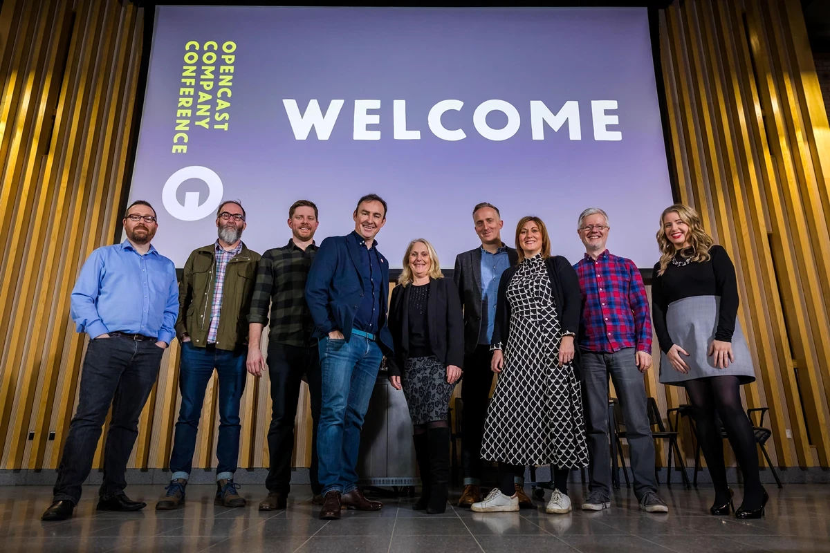A group of people stand on a stage in front of a large screen displaying the word ‘WELCOME’ at the Opencast Company Conference. The backdrop features vertical wooden panels and theatre-style lighting.