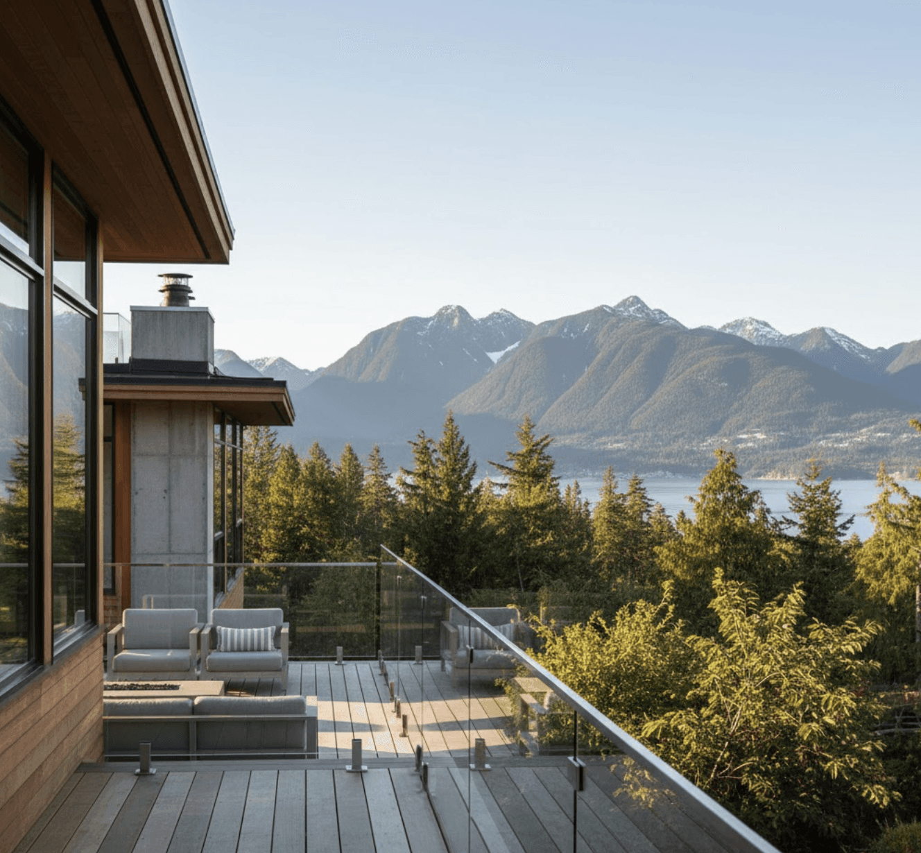 odern photorealistic image of frameless glass deck railing on West Coast contemporary home, background showing Vancouver North Shore mountains and coastal forest scenery, warm natural daylight, clean architectural lines emphasizing transparency, view through clear glass panels to mountain vista, professional architectural photography style, subtle blue-green coastal colour palette matching BC landscape, no text overlays, emphasis on unobstructed mountain view and sleek modern design, welcoming outdoor deck space