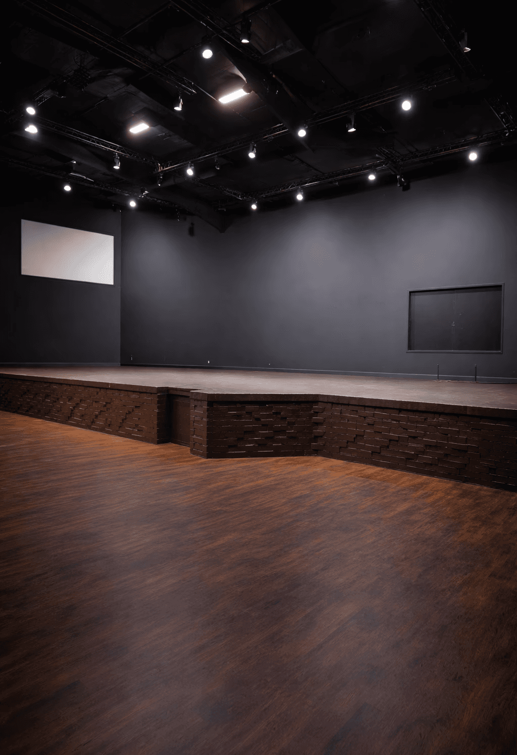 A modern, glass-walled house with warm interior lighting is perched on a rocky cliff overlooking a calm sea at sunset, under a clear blue sky.