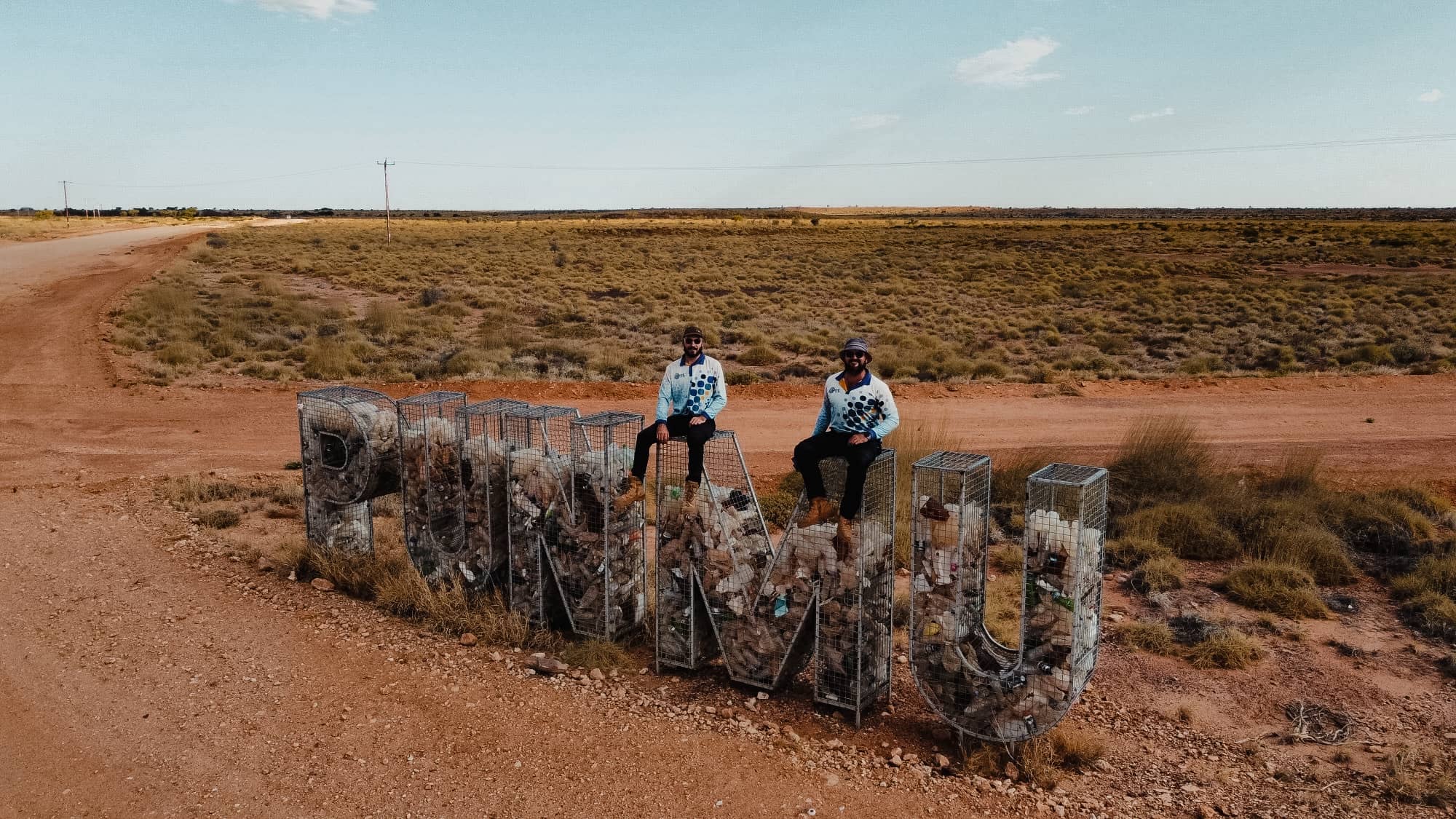 Two men sitting on a giant wire mesh sculpture filled with waste, spelling out "HOPE DOWNS" in a remote Australian landscape.