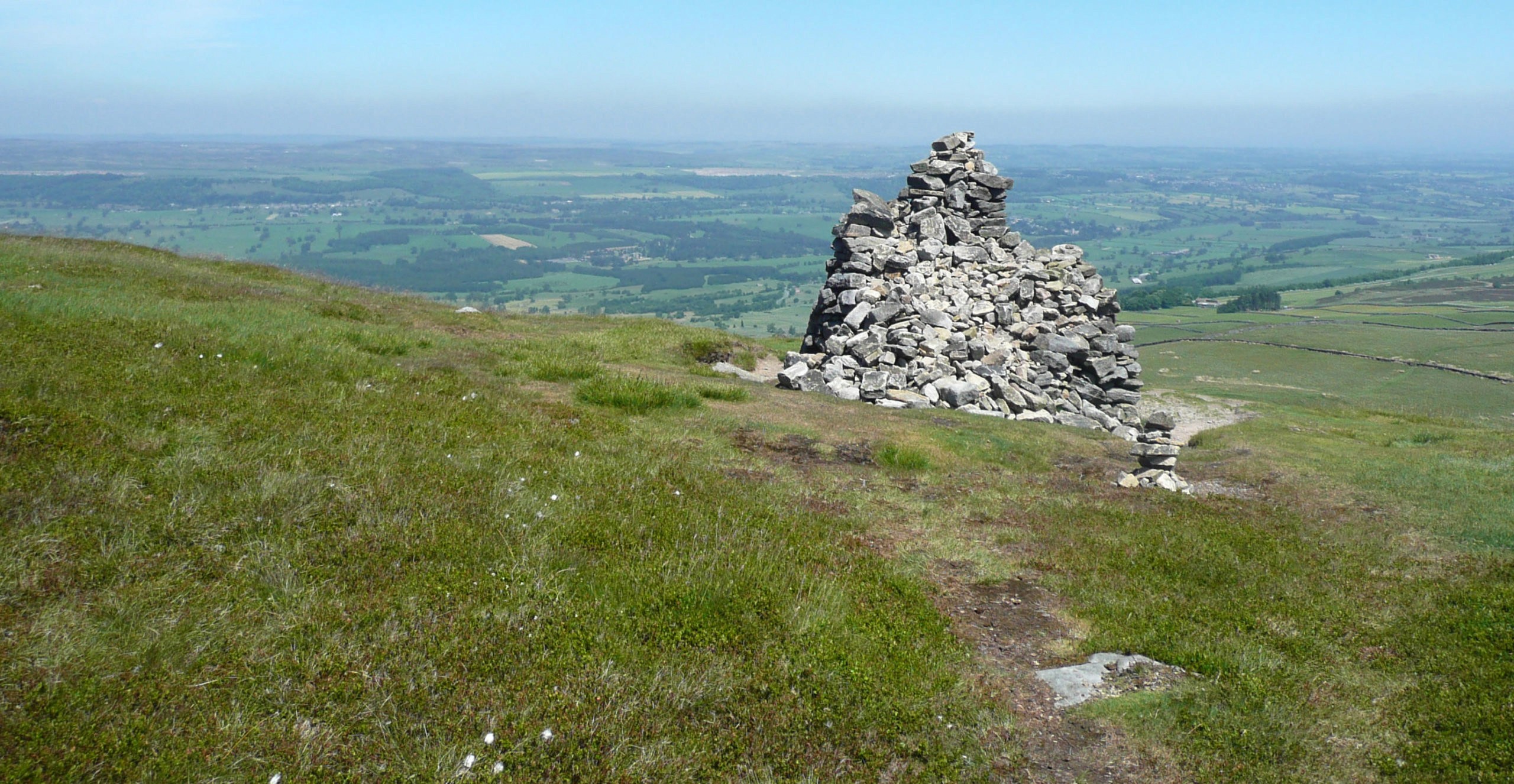 Stone cairn summit marker on grassy hilltop with panoramic views across sprawling countryside landscape under blue sky