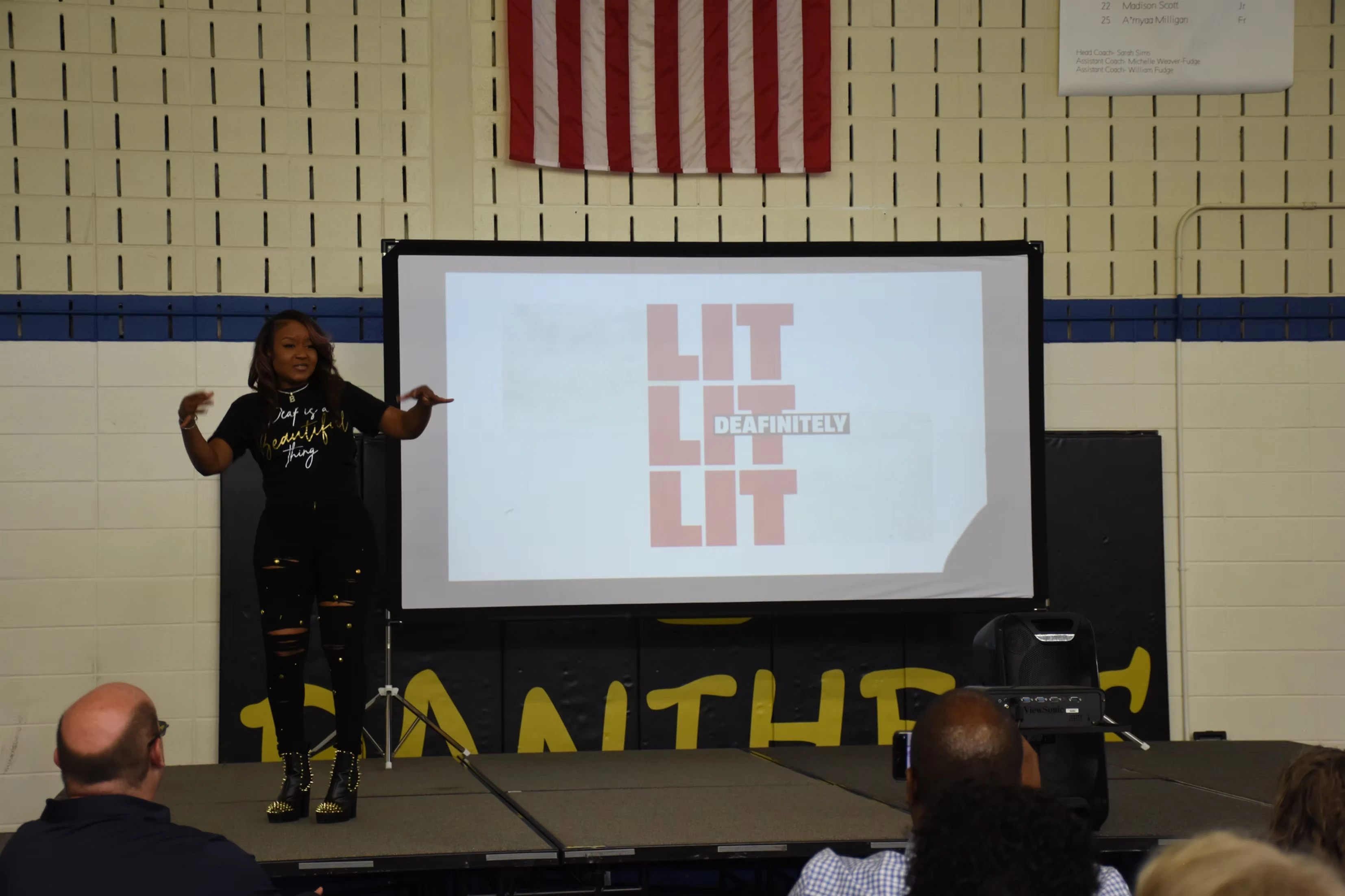 A woman giving a presentation on a stage. The presentation's title is "LIT" repeated three times. She is gesturing energetically.