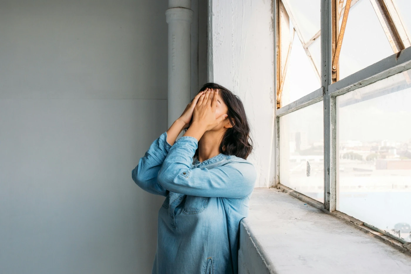 Eine Frau steht am Fenster eines minimalistischen Raumes und bedeckt ihr Gesicht mit den Händen. Das Bild symbolisiert emotionale Belastung, kognitive Überforderung und den Bedarf nach Klarheit und Entlastung.