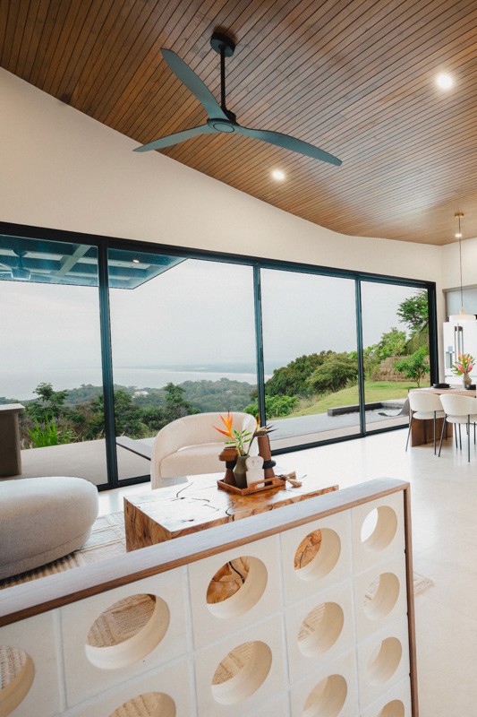 Open-concept living and dining area with floor-to-ceiling glass, wood ceiling, and panoramic ocean and jungle views.