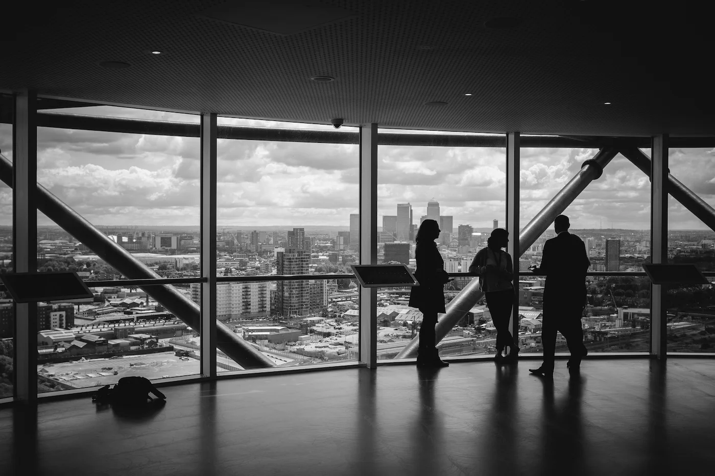 High-contrast black and white photograph showing the silhouette of three people standing inside an observation deck with floor-to-ceiling windows, looking out over a sprawling city skyline under a dramatic, cloudy sky.