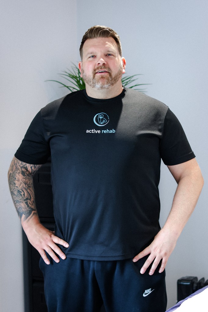 Close-up portrait of a person wearing a gray insulated hooded jacket with the hood up, showing their face framed by the hood against a light background.