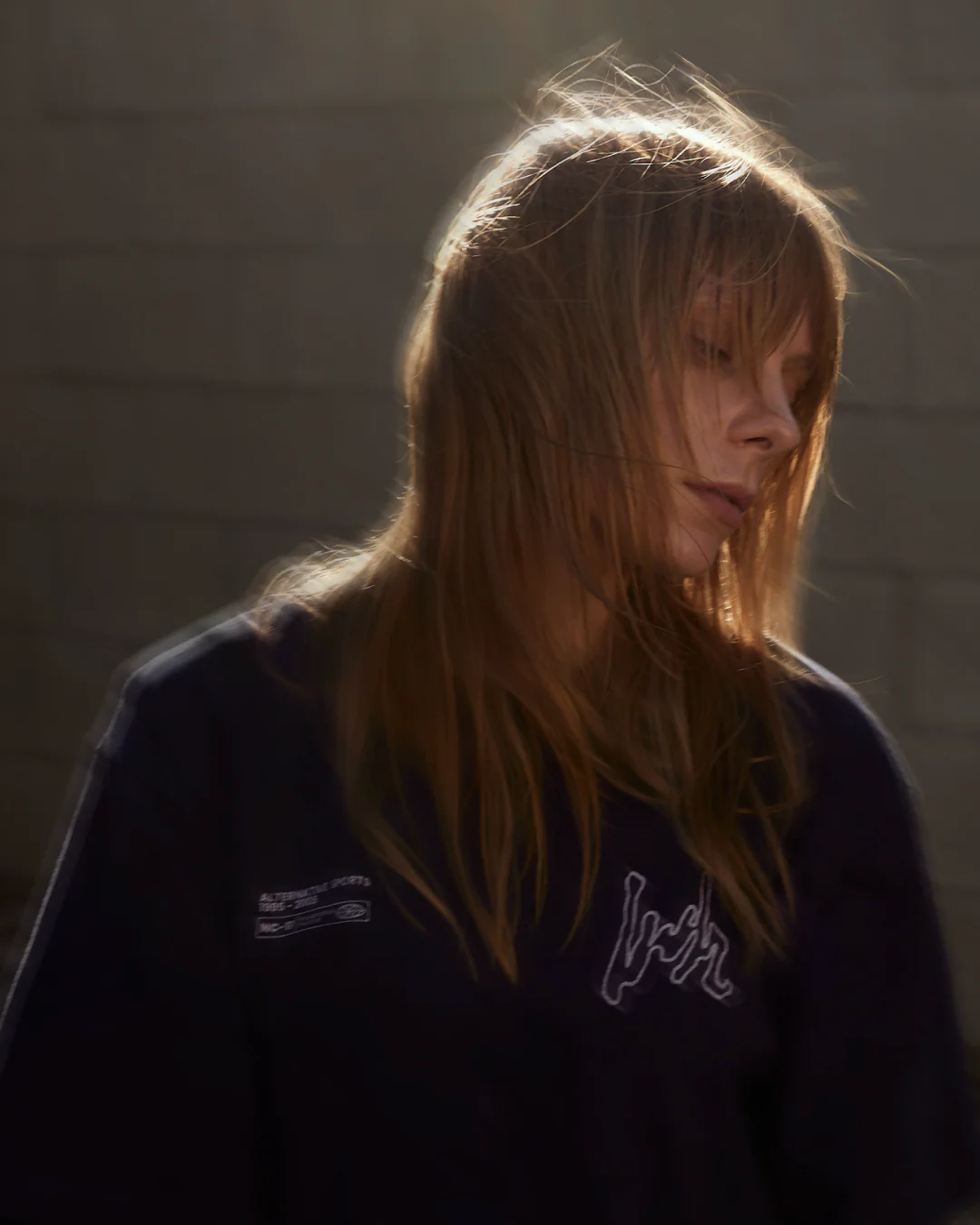 A woman in afternoon light with her hair blowing, wearing the Volume LWH Alternative Sports 02 T-shirt in front of a sunlit cinderblock wall.