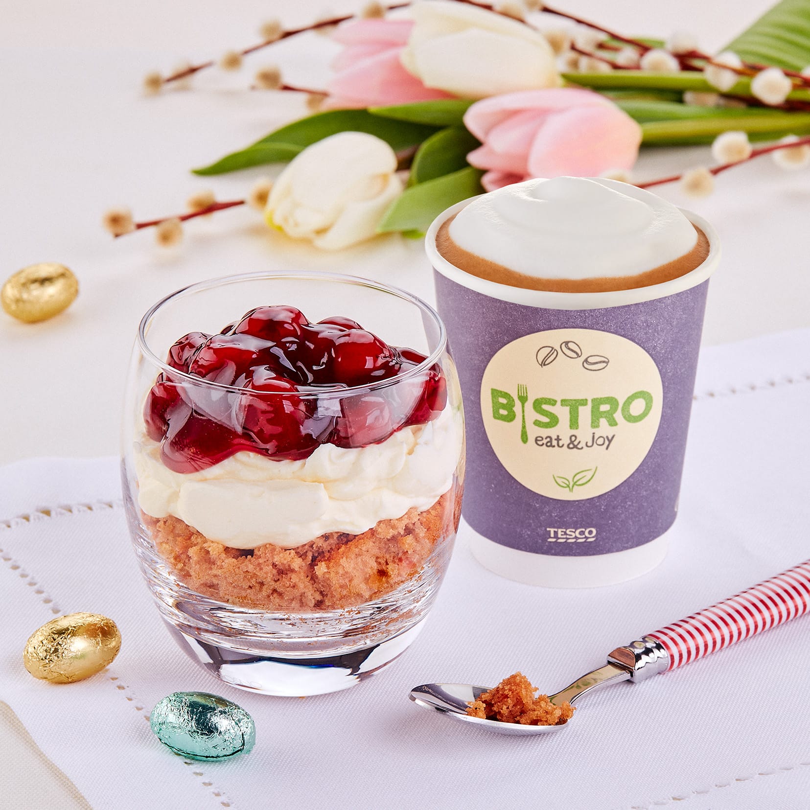 A dessert featuring a layered cherry parfait with whipped cream and a crumb base, accompanied by a take-out cup with "Tesco Bistro" branding, sits beautifully arranged on a white napkin, surrounded by ornamental eggs and tulips for a food photo session.