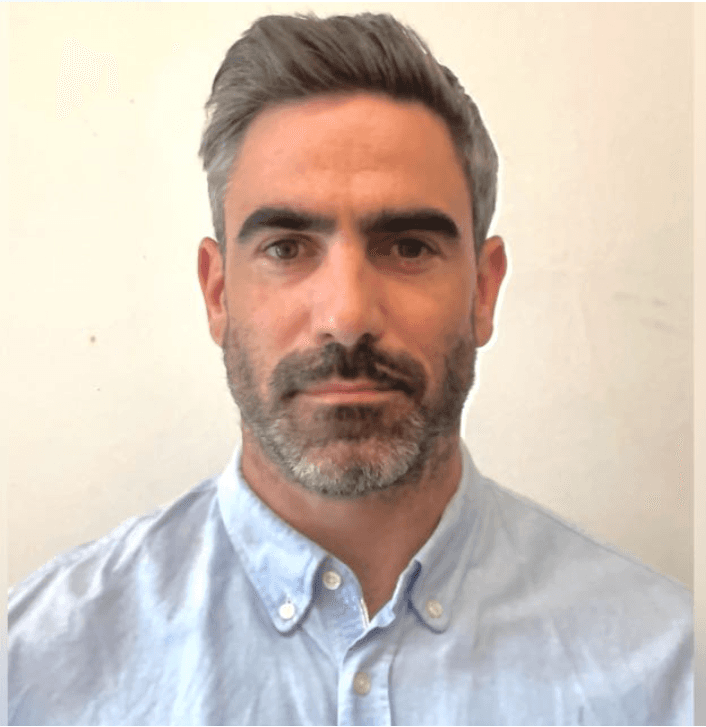 A young person with short, light-colored hair and glasses is looking directly at the camera.