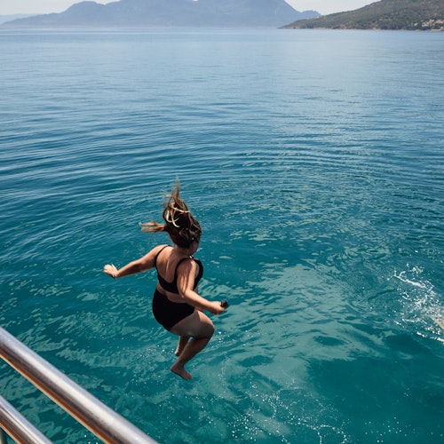 一個穿著黑色泳衣的人從船上跳入清澈的藍色海水中，背景是一些島嶼。