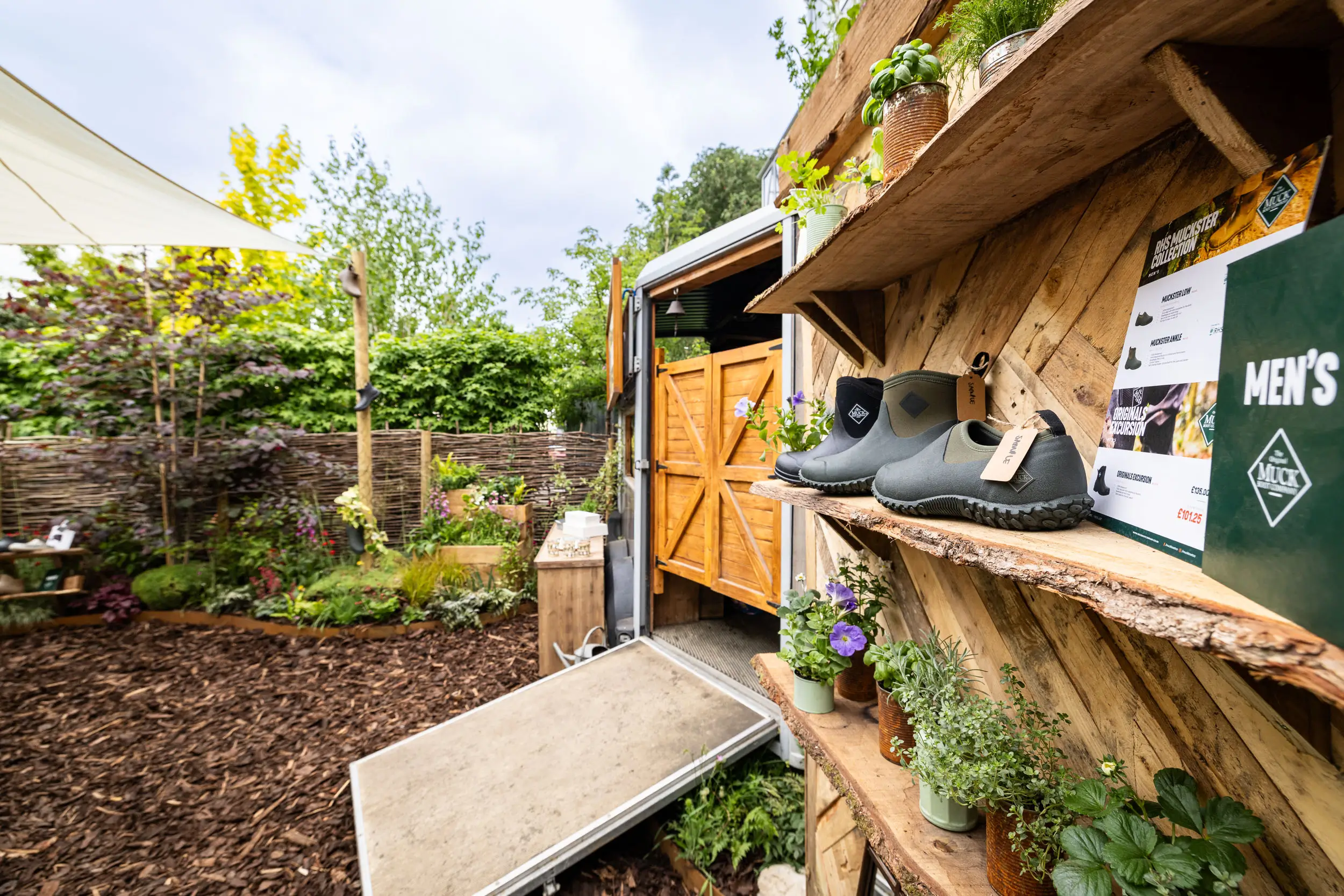 A colorful garden featuring wooden shelves with plants, surrounded by greenery and a light blue sky.