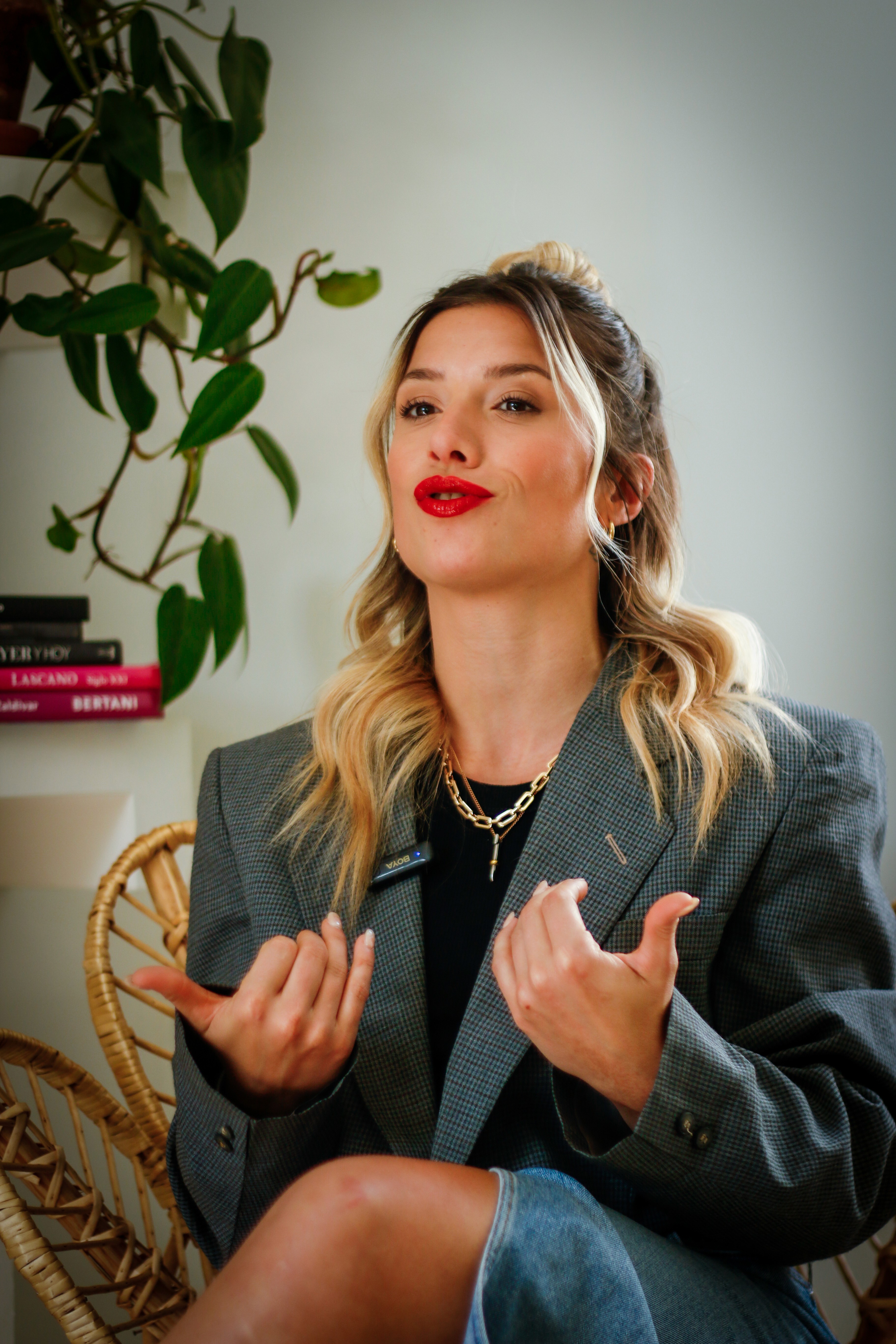 Mujer con blazer gris y labial rojo gesticula mientras habla; sentada en silla de mimbre, con planta y libros de fondo.