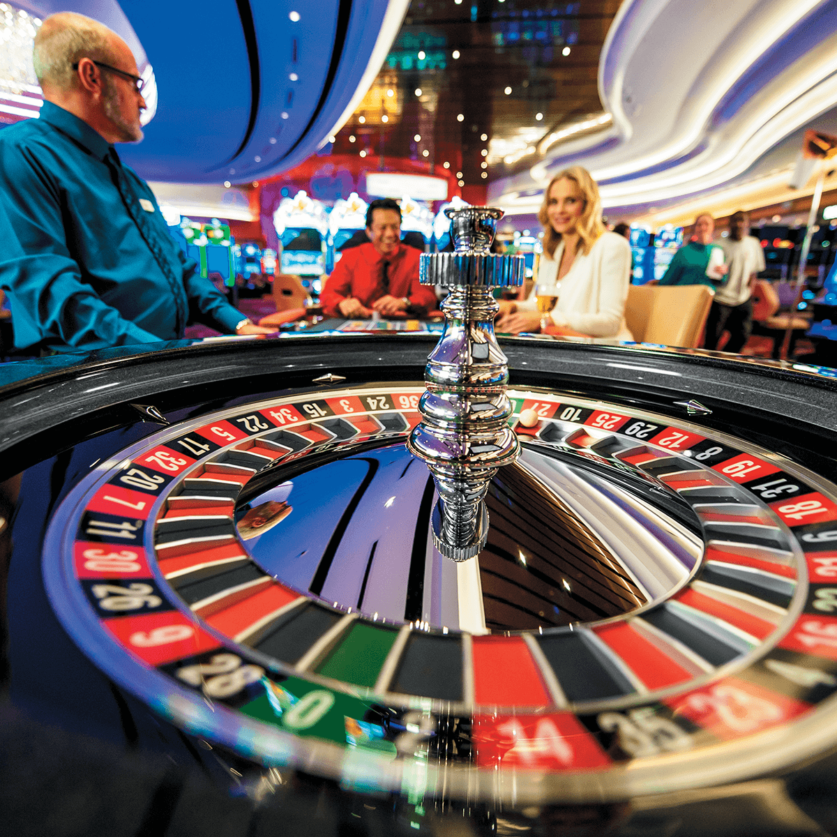 A close-up view of a roulette wheel in a casino with three people standing and sitting in the background, engaged in conversation.